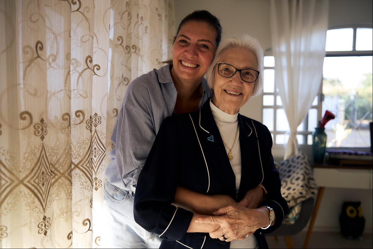 Una mujer sonriente está de pie detrás de otra que la rodea con los brazos junto a una ventana con cortinas en el interior de una casa