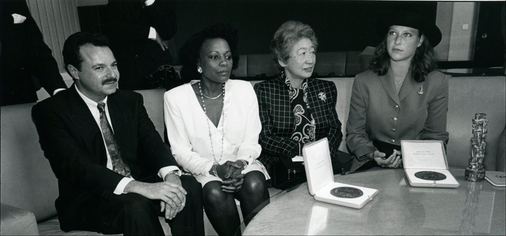 Un grupo de cuatro personas sentadas una junto a la otra (de izquierda a derecha, un hombre, una mujer afro, una mujer japonesa y una mujer noruega)