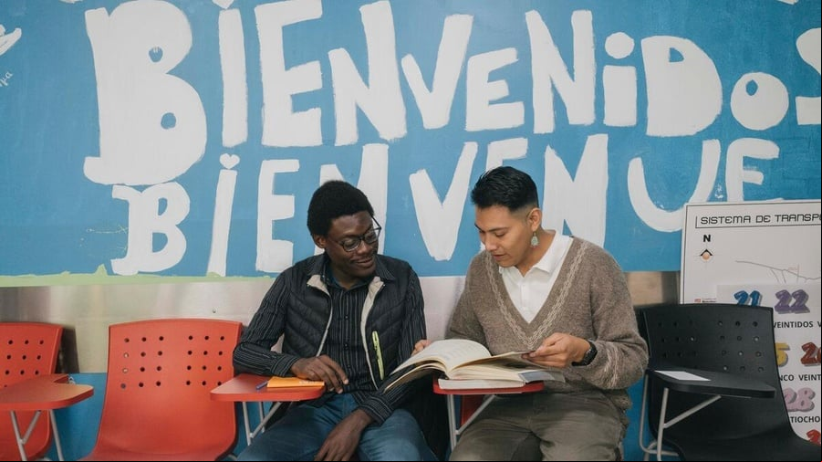 Dos hombres sentados en un salón de clases revisan un libro. Detrás de ellos se lee pintada en la pared la palabra Bienvenidos.